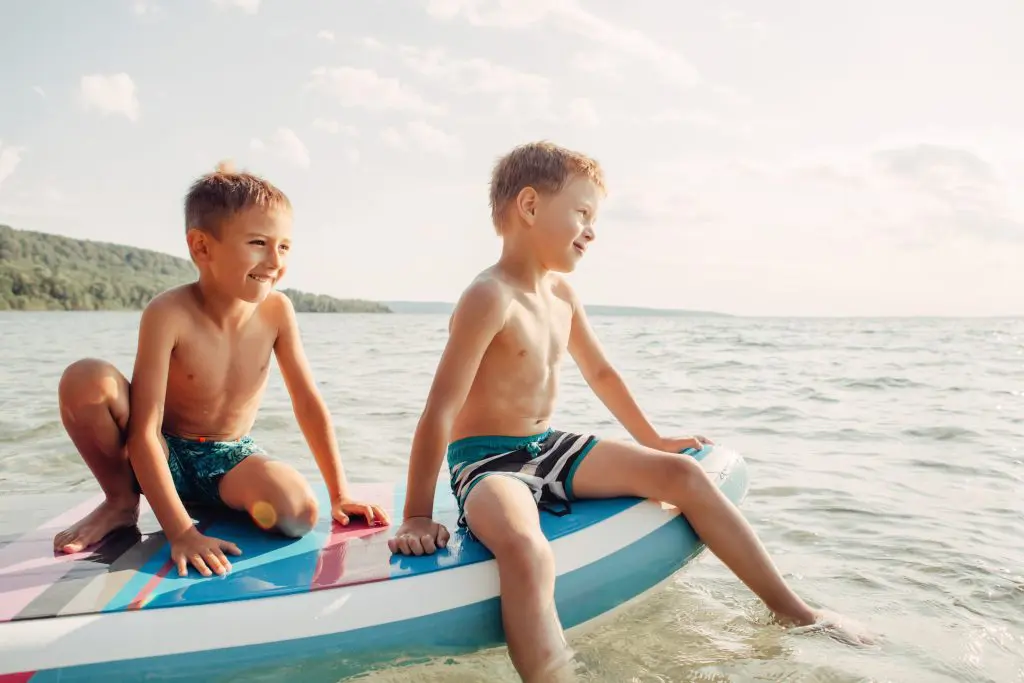 Two smiling Caucasian boys kids sitting on paddle sup surfboard