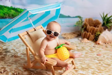 91 Summer Inspired Boy Names From Sun, Sea to Sand 1 Happy little boy in glasses, lies on a wooden deck chair, sunbathes on a sandy beach with palm trees by the sea and drinks juice under a beach umbrella
