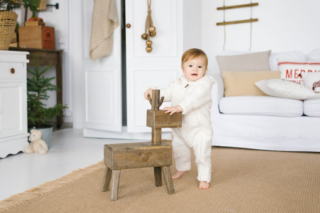 One-year-old child in a soft jumpsuit stands next to a wooden deer toy