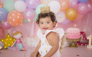 Cute baby girl with balloons and birthday cake sitting at home